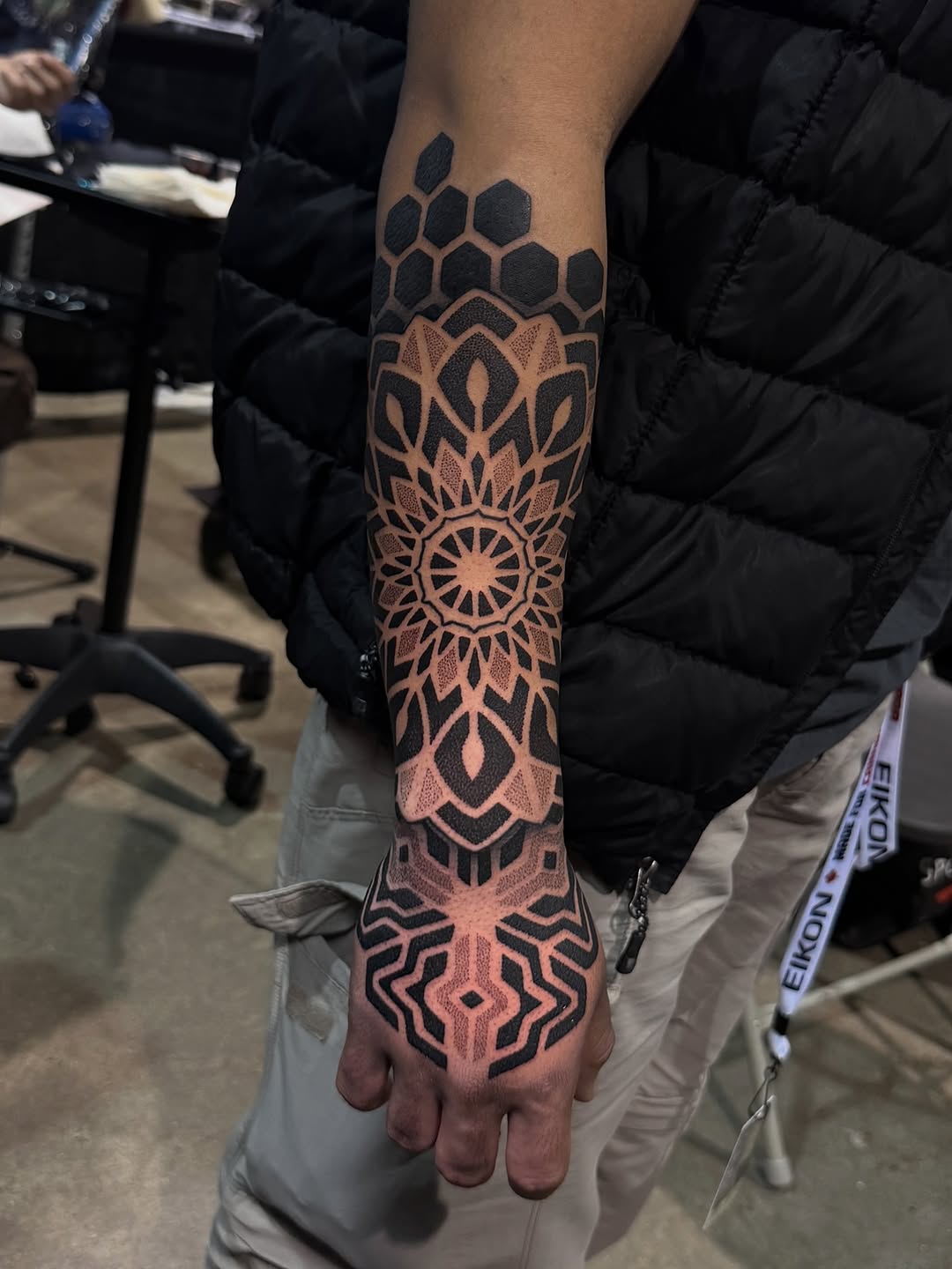 Symmetrical mandala and honeycomb pattern tattoo covering forearm and hand in black and natural skin tones.