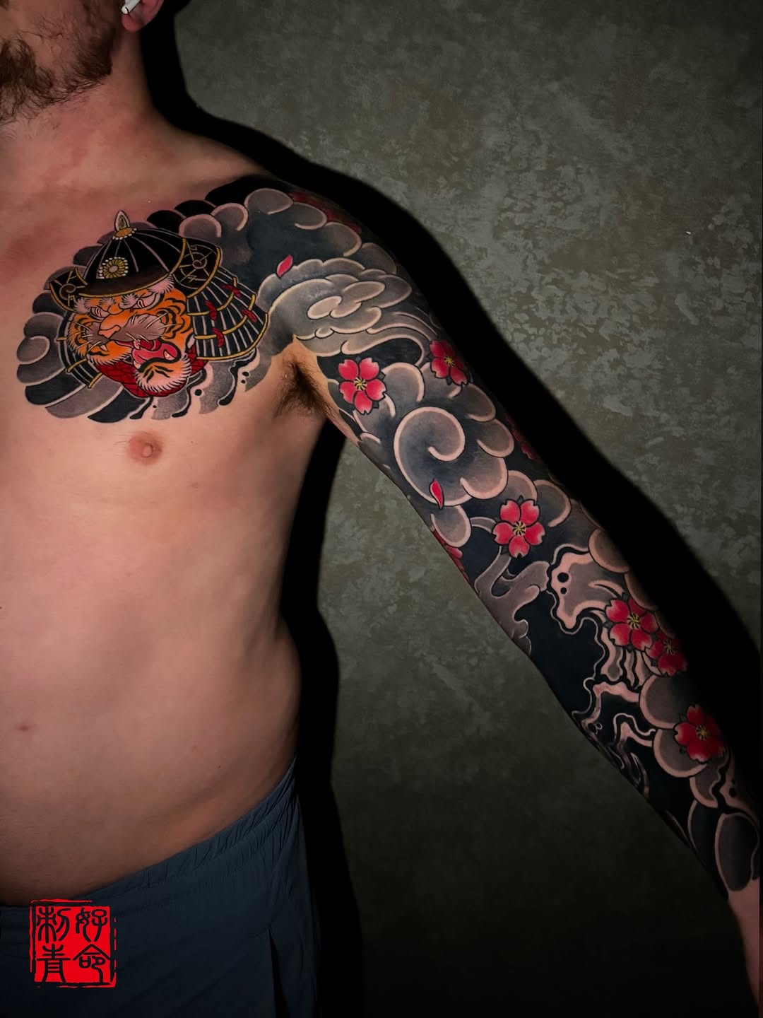 Japanese-inspired sleeve tattoo featuring an oni mask with helmet, ornamental clouds, and red flowers on chest and arm.